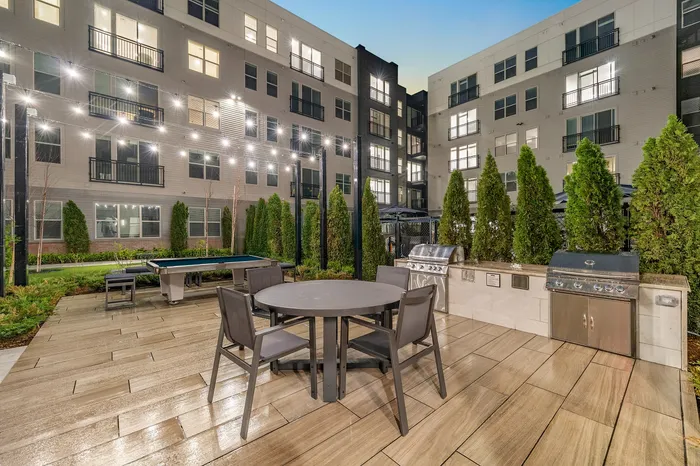 Modern outdoor communal area with elegant lighting, featuring a stylish dining setup, BBQ grills, and a lush garden setting in a contemporary apartment complex.