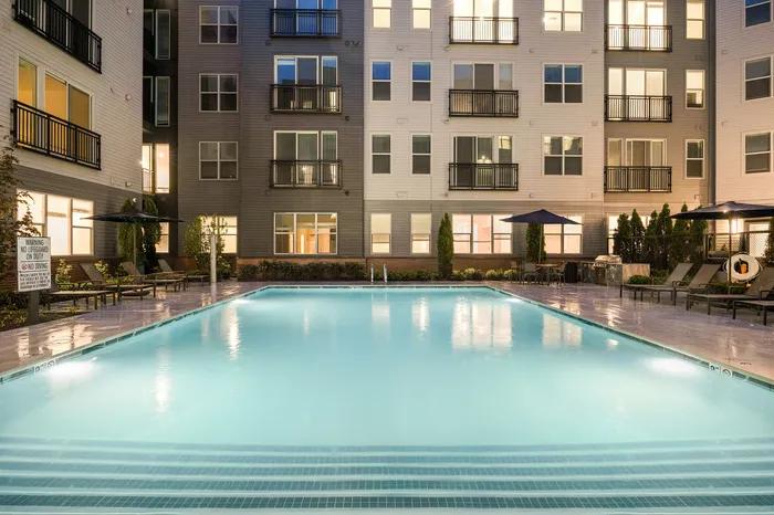 Luxurious apartment complex featuring a beautifully illuminated swimming pool surrounded by elegant lounge chairs and modern architecture.