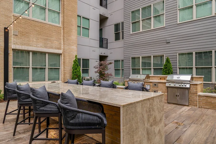 Elegant outdoor communal area featuring a modern dining setup with sleek bar stools, stylish granite countertop, and built-in stainless steel grills, surrounded by a sophisticated apartment building fa&ccedil;ade.