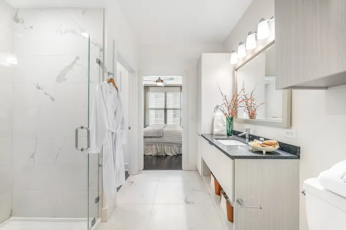 Elegant bathroom featuring a luxurious shower, plush white robe, and modern vanity with stylish decor, leading to a cozy bedroom with natural light.