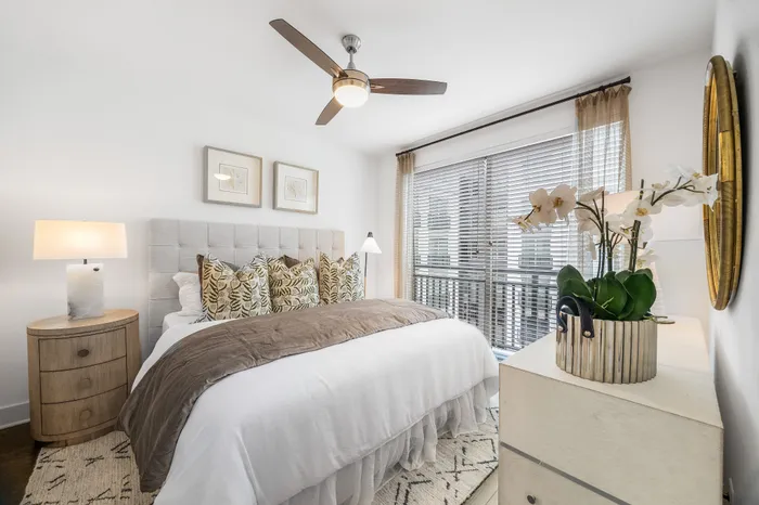 Bright and elegant bedroom featuring a plush bed with patterned pillows, modern ceiling fan, and expansive window with stylish blinds.