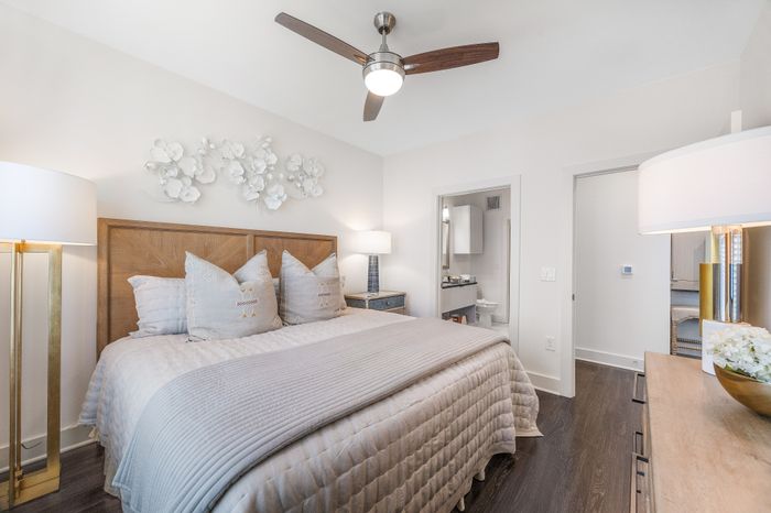Bright and elegant bedroom featuring a plush king-sized bed with stylish decorative pillows, modern ceiling fan, chic wall art, and a glimpse into the en-suite bathroom.