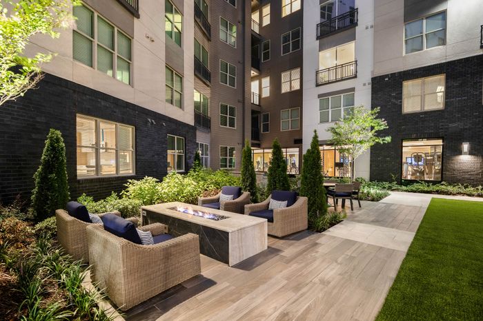 Elegant courtyard in a modern apartment complex featuring cozy seating around a sleek outdoor fire pit, surrounded by lush greenery and well-lit windows.