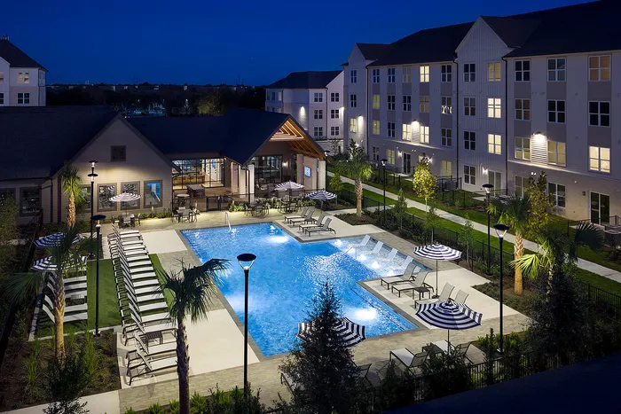 Elegant nighttime view of apartment complex pool area, featuring illuminated swimming pool, comfortable lounge chairs, and stylish umbrellas surrounded by landscaped gardens.