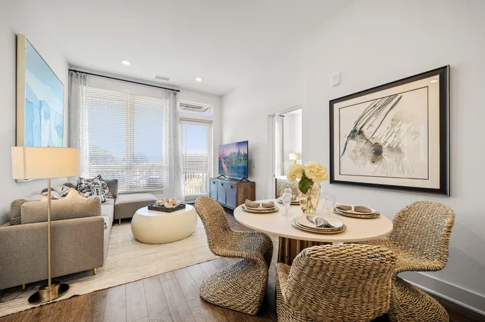 Bright and inviting living and dining area featuring a cozy sofa, stylish decor, and a round dining table with woven chairs, complemented by large windows letting in natural light.