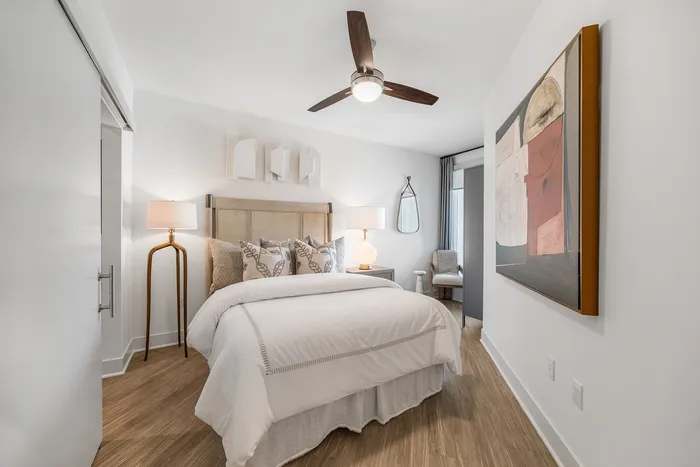 Elegant modern bedroom with stylish decor, featuring a plush bed with decorative pillows, artistic wall art, and a cozy sitting area. Bright and spacious with natural wood flooring.