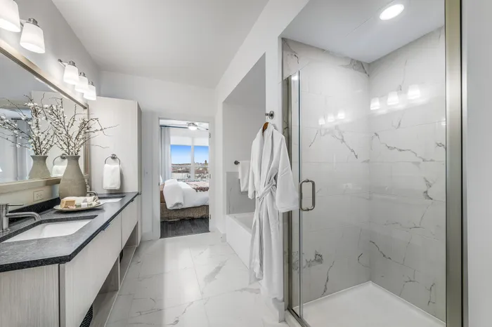 Luxurious bathroom with modern fixtures, featuring a sleek glass-enclosed shower, elegant marble tiles, and a view into a cozy bedroom with cityscape views.