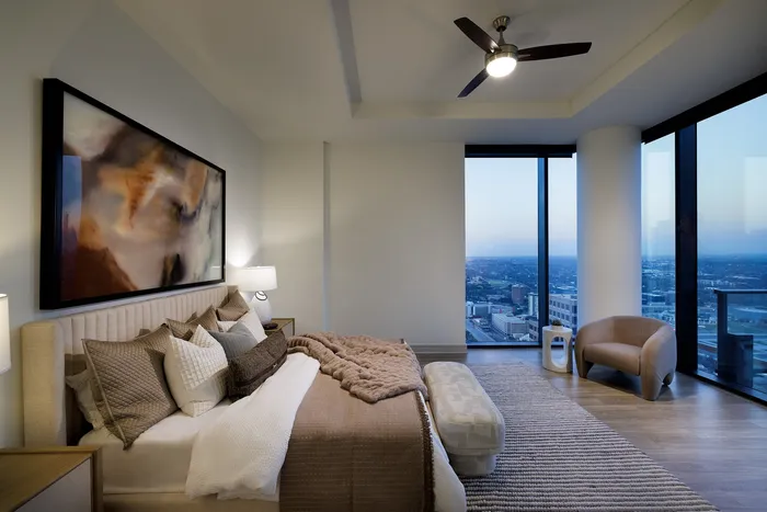 Luxurious bedroom with floor-to-ceiling windows, offering breathtaking city views. Elegant decor featuring a plush bed, modern art, and cozy seating area.