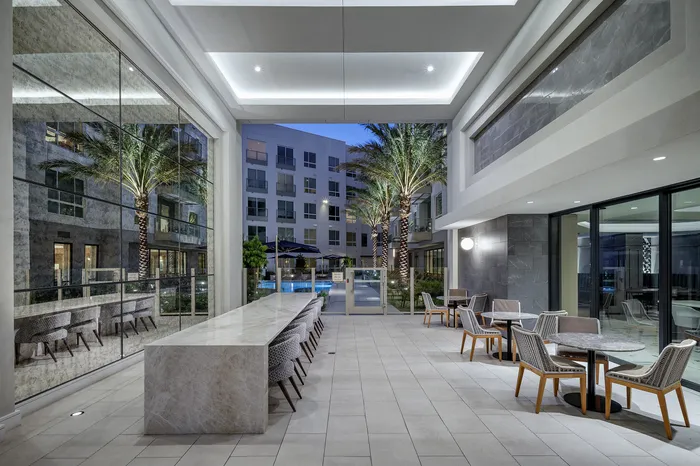 Elegant communal lounge featuring modern marble dining table, stylish seating, and open view of illuminated courtyard with palm trees and pool.