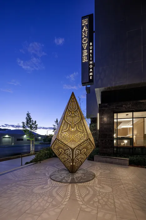 Elegant evening view of Hanover Burlingame with an intricate, illuminated sculpture casting captivating shadows on the walkway.