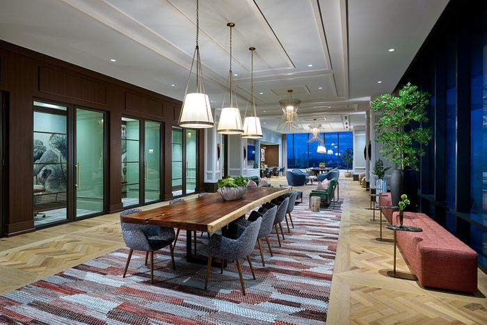 Elegant dining and lounge area with modern lighting, large wooden table, plush seating, and floor-to-ceiling windows showcasing a stunning city view.