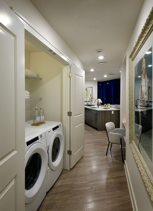 Modern apartment hallway featuring a sleek laundry area with washer and dryer, stylish wood flooring, and an elegant kitchen in the background.