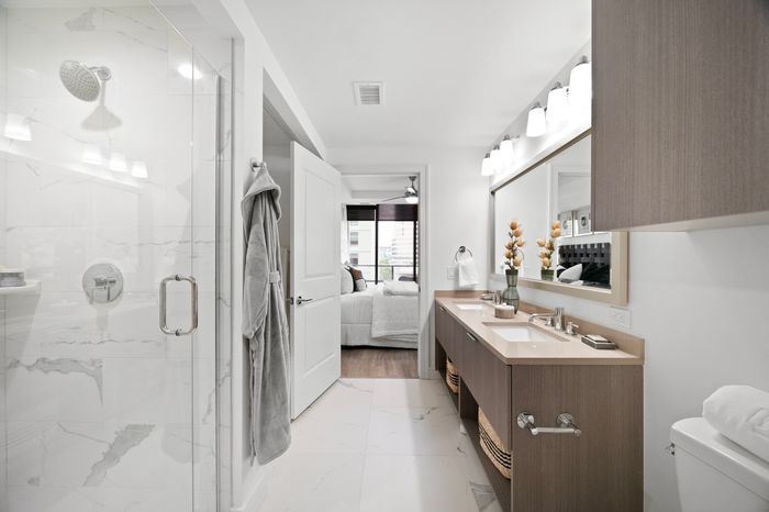 Elegant modern bathroom with dual sinks, sleek wood cabinetry, and a spacious shower, leading into a bright bedroom with a large window.