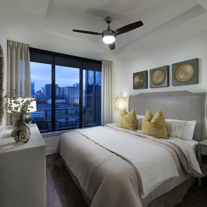 Elegant bedroom with city skyline view, featuring a plush bed adorned with decorative pillows and modern art on the walls, accentuated by warm lighting and contemporary furnishings.