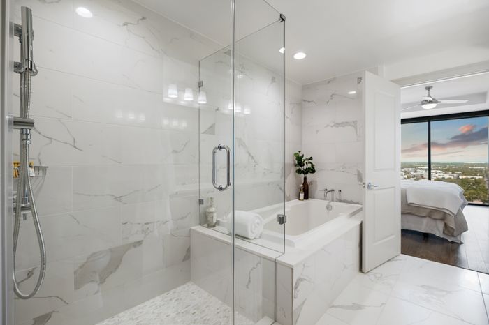 Luxurious bathroom with elegant marble tiles, glass-enclosed shower, and soaking tub, leading to a bedroom with panoramic city views.