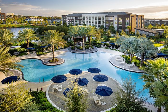 Luxurious apartment complex featuring a sprawling resort-style pool with palm trees, stylish lounge chairs, and charming blue umbrellas, set against a backdrop of lush greenery and modern architecture.