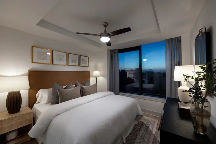 Cozy bedroom at dusk with elegant decor, featuring a large bed, modern ceiling fan, ambient lighting, and a stunning city view.