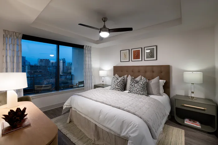 Elegant bedroom with stylish decor, featuring a plush bed with patterned cushions and views of a cityscape through a large window.