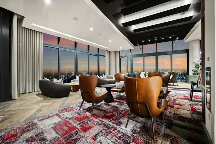 Luxurious living room with panoramic city views, featuring elegant leather chairs, modern decor, and a vibrant patterned rug, bathed in sunset hues.