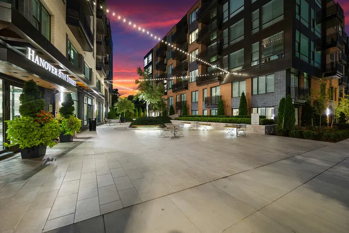 Charming courtyard of a modern apartment complex with string lights glowing against a vibrant sunset sky, surrounded by lush greenery and contemporary architecture.