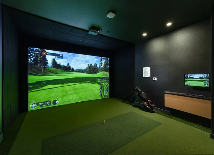 Modern apartment featuring an indoor golf simulator room with a large screen displaying a picturesque golf course, plush green turf, and a golf bag, perfect for entertainment and relaxation.