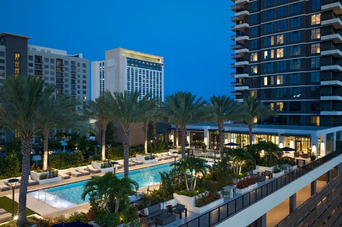 Luxurious high-rise apartment complex with palm trees surrounding a sparkling pool and stylish outdoor seating, beautifully illuminated at dusk.