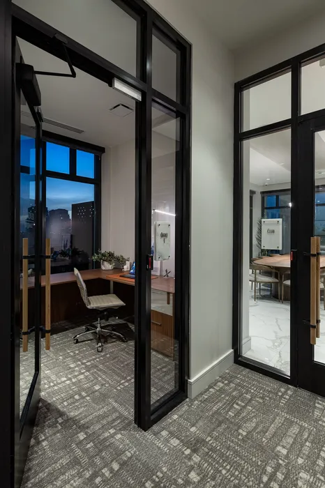 Modern office space with glass doors, stylish grey carpet, and a sleek desk overlooking cityscape.