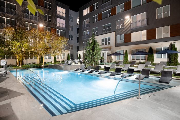 Luxurious apartment complex featuring a beautifully illuminated swimming pool, surrounded by elegant lounge chairs and lush greenery, creating a perfect oasis for relaxation.