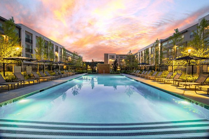 Luxurious apartment complex featuring a stunning outdoor pool surrounded by comfortable lounge chairs and beautifully landscaped gardens, set against a breathtaking sunset sky.