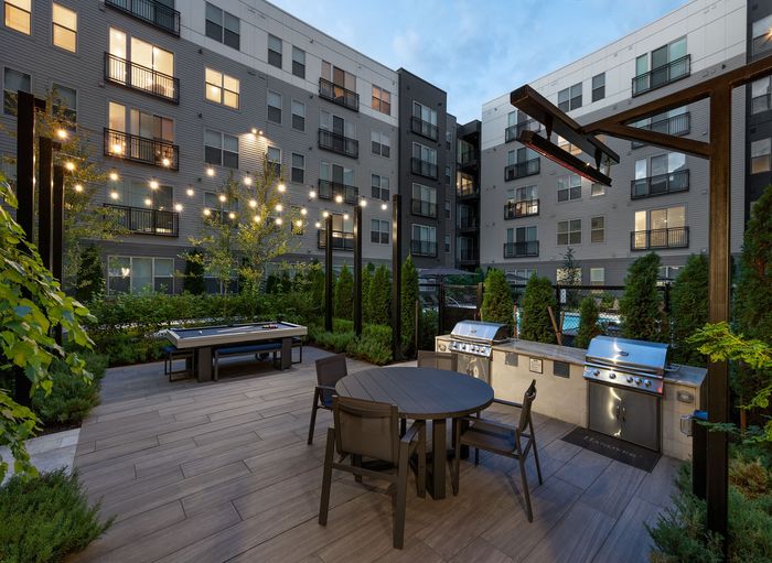 Chic urban patio with stylish seating, barbecue grills, and ambient string lights, surrounded by lush greenery against a modern apartment backdrop.