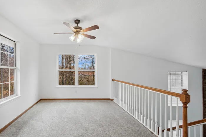 Sunlit loft area with carpeted floor, large windows, and a ceiling fan.