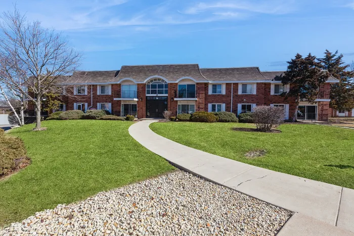Charming brick apartment building with manicured green lawns and a welcoming pathway under a clear blue sky.