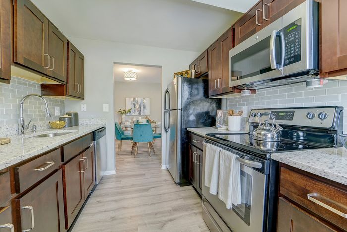 Chic modern kitchen at ReNew White Marsh with sleek stainless steel appliances, granite countertops, and rich wooden cabinets, leading to a vibrant dining area with stylish teal chairs.