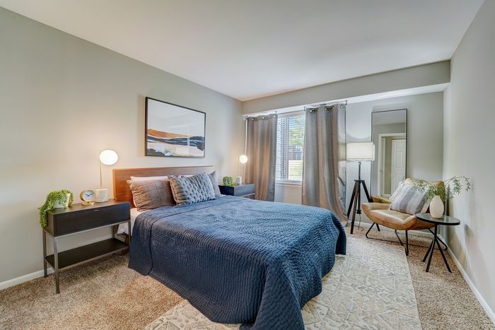 Inviting bedroom at ReNew White Marsh with modern decor, featuring a cozy bed with plush pillows, stylish nightstands, and elegant lighting, complemented by a large window with curtains and a comfortable reading chair.