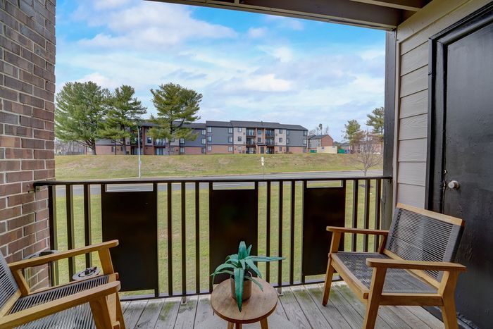 Charming apartment balcony at ReNew White Marsh with cozy wooden seating, vibrant plant, and serene view of lush greenery and modern building.