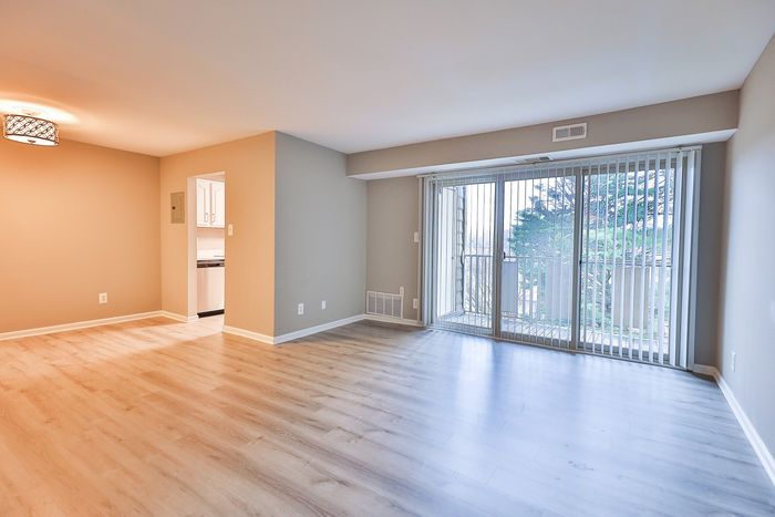 Bright and spacious living room at ReNew White Marsh with sleek wooden flooring, large windows with vertical blinds, and a modern kitchen entrance.