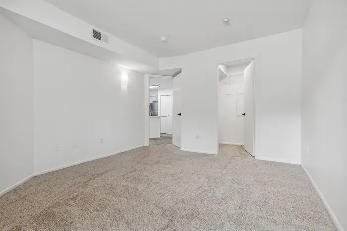 Bright and inviting bedroom with plush carpet and modern white walls, leading to a spacious walk-in closet and kitchen entrance.