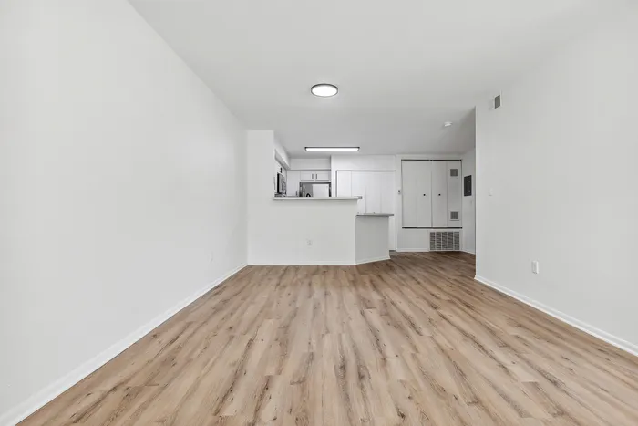 Bright open-concept living area with elegant wood flooring leading to a modern, fully equipped kitchen. Sleek white cabinetry and stainless steel appliances enhance the inviting and spacious ambiance.