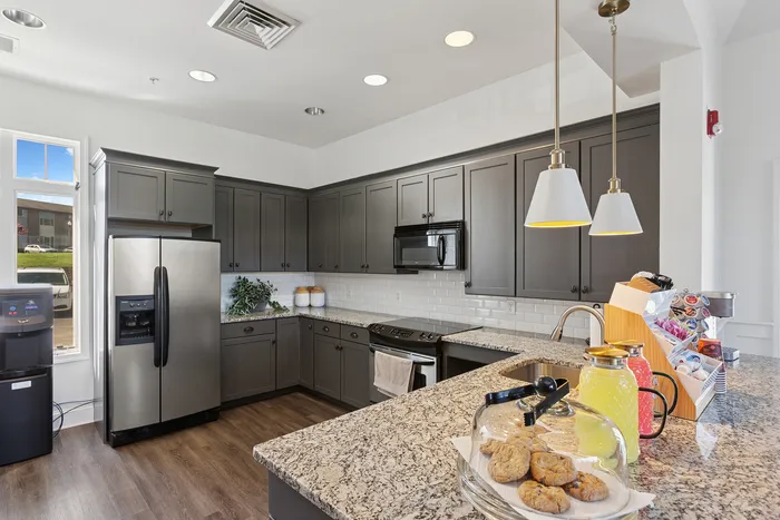 Modern kitchen at ReNew Howard Crossing with sleek gray cabinets, stainless steel appliances, and elegant pendant lighting. Features a granite countertop island with refreshments and cookies, creating a warm and inviting space.