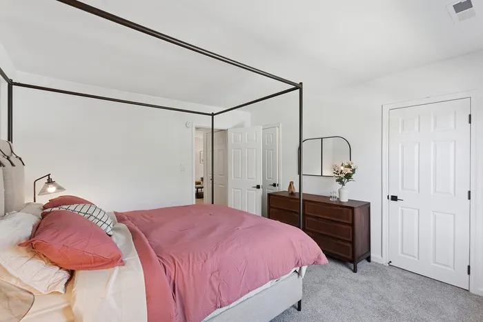Elegant bedroom at ReNew Howard Crossing featuring a cozy four-poster bed with pink bedding, a wooden dresser with a stylish mirror, and soft carpeted flooring.