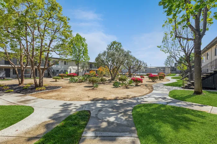 Inviting apartment courtyard showcasing a landscaped garden with vibrant flowers, winding paths, and lush green trees under a clear blue sky.