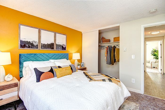 Vibrant bedroom featuring a plush bed with colorful pillows, striking orange accent wall, and open closet space at ReNew Dash Point.