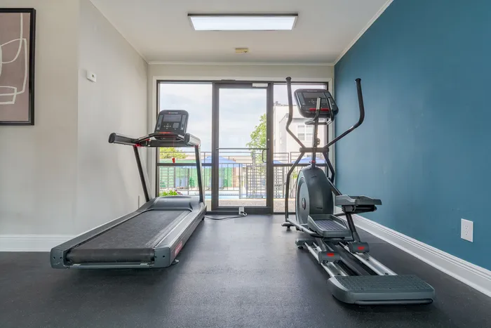 Bright and modern gym room featuring premium treadmill and elliptical machine with a view of the outdoor pool area, enhanced by sleek blue accent wall.