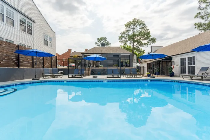 Beautiful apartment complex pool area with inviting blue water, surrounded by lounge chairs and vibrant blue umbrellas, perfect for relaxing under the sunny sky.