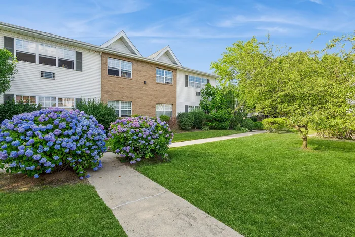 Charming apartment building surrounded by lush greenery and vibrant hydrangeas, featuring a well-maintained pathway and crisp exterior.