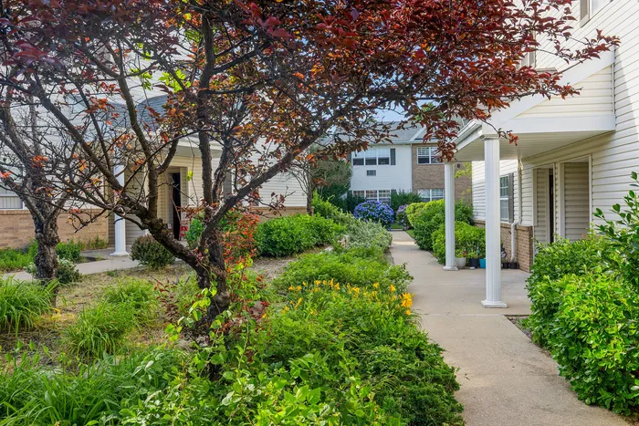 Charming apartment exterior with lush gardens and a welcoming walkway shaded by colorful trees.