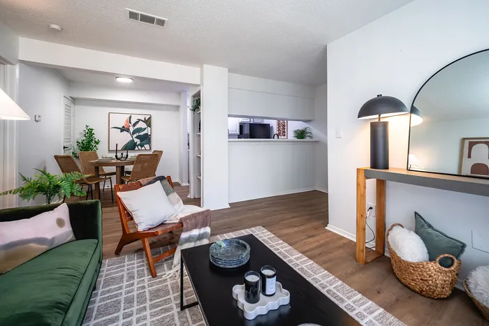 Chic apartment interior featuring a cozy living space with a modern green sofa, stylish wooden chair, elegant floor lamp, and a sleek console table. The adjacent dining area showcases a warm, inviting atmosphere with wicker chairs and a vibrant plant-themed artwork.