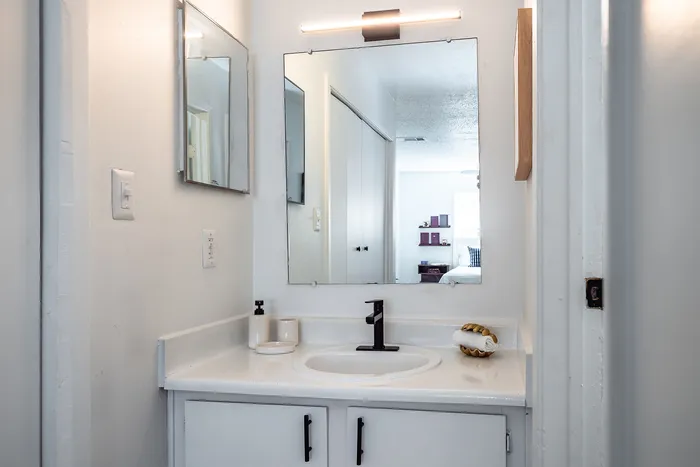 Modern bathroom vanity with sleek black fixtures, ample mirror space, and elegant lighting, showcasing a peek into a cozy bedroom.
