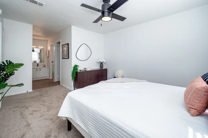 Bright and airy bedroom with elegant decor, featuring a modern ceiling fan, plush carpeting, and a stylish vanity leading to an ensuite bathroom.