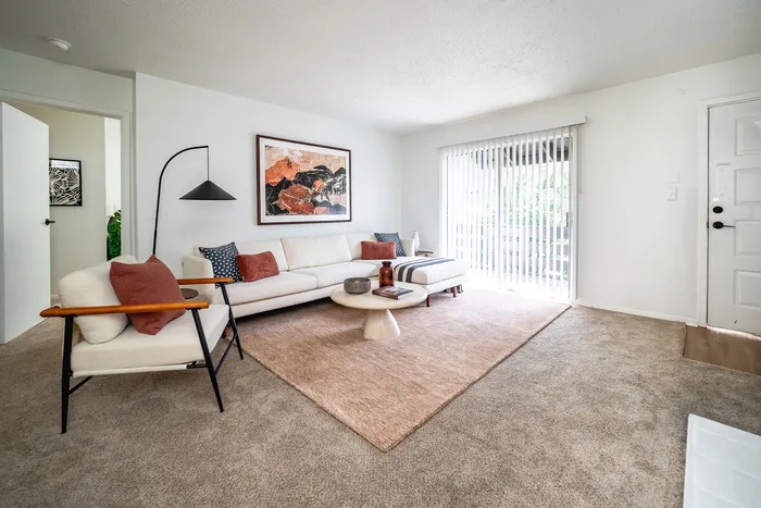 Bright and inviting living room showcasing modern beige furniture, contemporary art on the wall, and a view through sliding glass doors to a serene outdoor space.
